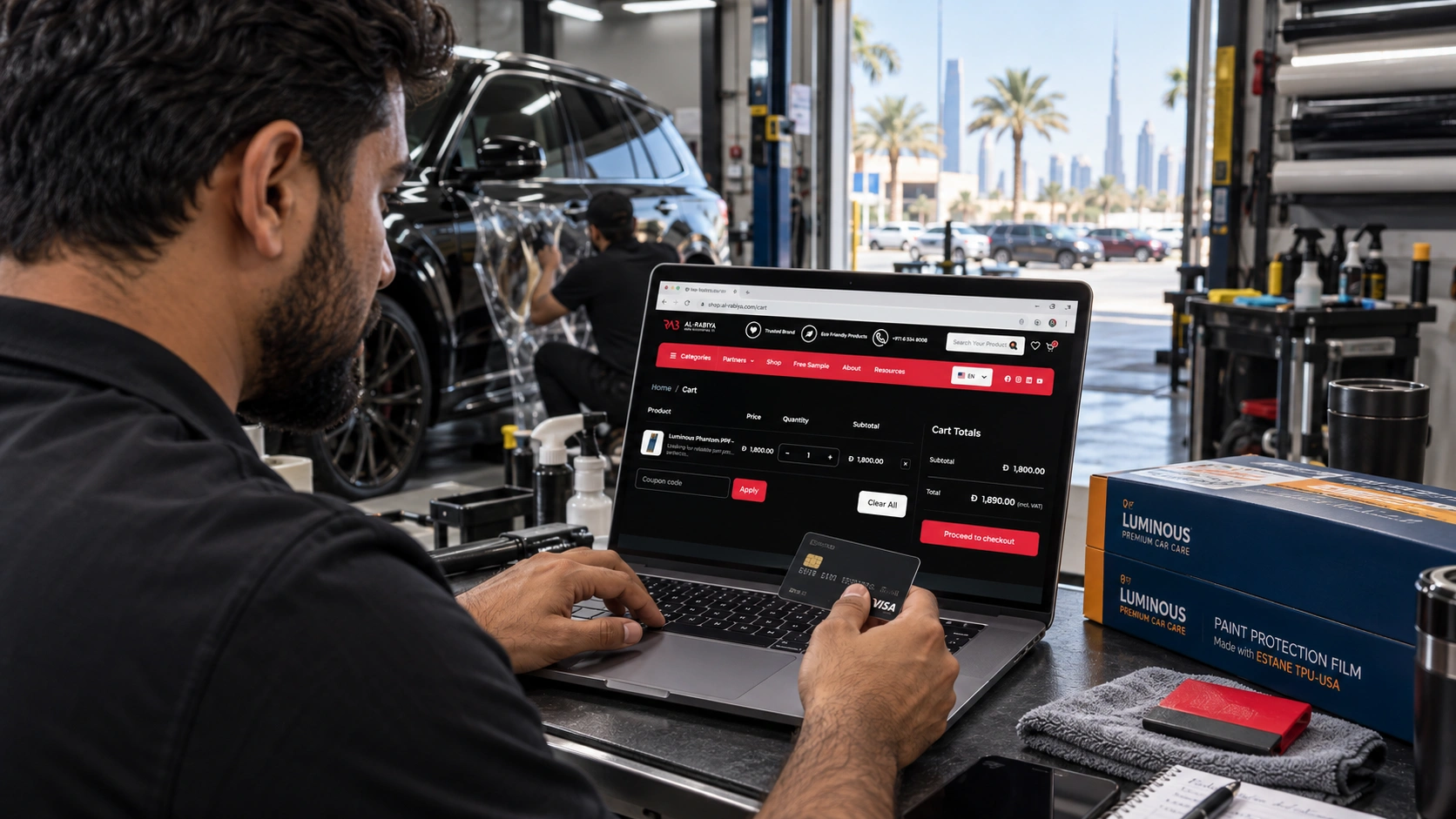 Installer is holding a physical bank/credit card in one hand, preparing to complete payment for an order on the Al-Rabiya automotive e-commerce UAE website displayed on the laptop.